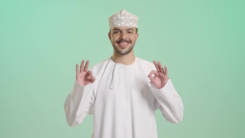 نظرات الحماس والشعور بالرضا, الموافقة والقبول والنصيحة، التعبير عن الجودة والاتقان، بورتريه لشاب عربي خليجي عماني يرتدي الدشداشة و الكمة يرفع يديه بإيماءات الجودة والامتياز والتميز، إيجاد الأفكار والحلول المناسبة, خلفية خضراء
