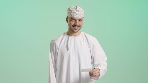 الشراء والدفع عن طريق بطاقات الإئتمان البنكية، إمكانية تسديد الفواتير عبر بطاقة الصراف, بورتريه لشاب عربي خليجي عماني يرتدي الدشداشة و الكمة  يمسك بيده البطاقة الالكترونية ينظر الى الكاميرا بإيماءات السرور, خلفية خضراء