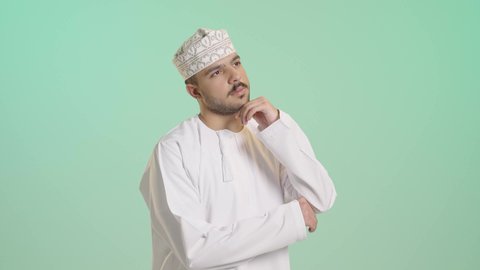 Concentration and focus to find a solution to a problem, a close-up portrait of an Arab Gulf Omani man wearing a dishdasha and kumma, resting his hand on his chin with expressions of thinking and concentration on something, gestures of distraction, confusion, and anxiety, contemplation and planning for the future, with a green background.