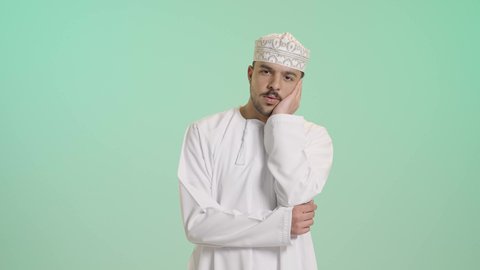 The feeling of boredom and annoyance, a portrait of an Arab Gulf Omani man wearing a dishdasha and kumma, resting his hand on his face, looking at the camera with gestures of focus and distraction, problem-solving and decision-making, green background.