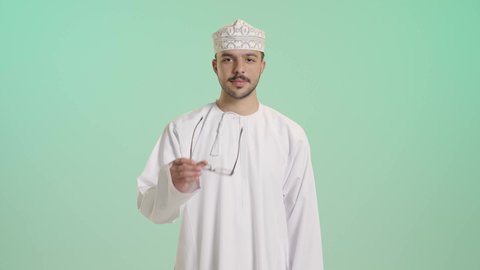 تحسين الرؤية ومحاولة تعديل البصر, ارتداء النظارات الخاصة بضعف النظر، الاهتمام بالمظهر الخارجي، بورتريه لشاب عربي خليجي عماني يرتدي الدشداشة و الكمة يضع النظارة الطبية، النظر الى الكاميرا بإيماءات السعادة، خلفية خضراء
