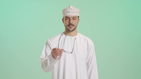 ارتداء النظارات الخاصة بضعف النظر، تحسين الرؤية ومحاولة تعديل البصر، الاهتمام بالمظهر الخارجي، بورتريه لشاب عربي خليجي عماني يرتدي الدشداشة و الكمة يضع النظارة الطبية، النظر الى الكاميرا بإيماءات السعادة، خلفية خضراء