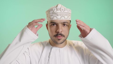 الاهتمام بالأناقة والمظهر الخارجي, بورتريه مقرب لشاب عربي خليجي عماني يرتدي الدشداشة والكمة يعدل هندامه بإيماءات تركيز, مفهوم الجاذبية والثقة بالنفس،خلفية خضراء