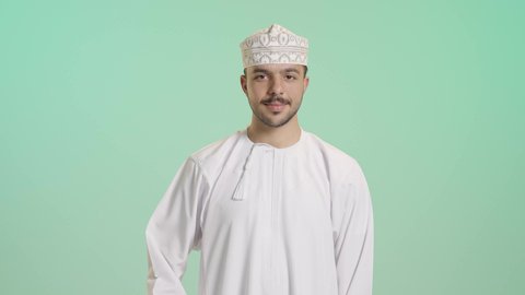 مفهوم تبادل المعلومات، حركة يد تدل على الاتصال، الاهتمام بالمظهر الخارجي والوسامة، بورتريه لرجل عربي خليجي عماني يرتدي الدشداشة والعمامة يرفع يده بايماءات الرد على المكالمات الهاتفية، تلقي أخبار سارة عبر الجوال، خلفية خضراء