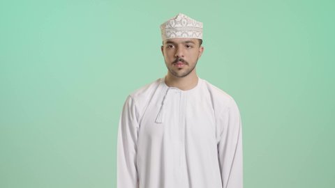 الشغف بمتابعة فريق كرة القدم, النظر إلى الكاميرا بإيماءات السعادة، بورتريه لرجل عربي خليجي عماني يرتدي الدشداشه و الكمة يتابع التفازبتعابير وجه ويدين تدل على الحماس والتشجيع، الانتصار وتحقيق الأهداف وفوز الفريق المفضل، خلفية خضراء