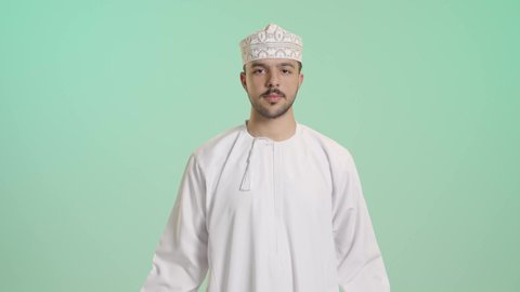 التمتع بالبنية الجسدية القوية, اللياقة البدنية والجسم الصحي المتين، بورتريه مقرب لرجل عربي خليجي عماني يرتدي الدشداشة والكمه يقوم بعرض عضلات يديه ينظر الى الكاميرا بإيماءات القوة، خلفية خضراء