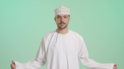 اللياقة البدنية والجسم الصحي المتين، بورتريه مقرب لرجل عربي خليجي عماني يرتدي الدشداشة والكمه يقوم بعرض عضلات يديه ينظر الى الكاميرا بإيماءات القوة، التمتع بالبنية الجسدية القوية، خلفية خضراء