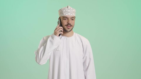 ايماءات السعادة و السرور، التواصل مع الاهل و الاصدقاء, بورتريه مقرب لشاب عربي خليجي عماني يرتدي الدشداشه و الكمه يقوم بإجراء مكالمة هاتفية عبر الهاتف المحمول، استخدام التقنيات الحديثة المتطورة, خلفية خضراء
