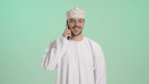 ايماءات السعادة و السرور، التواصل مع الاهل و الاصدقاء, بورتريه مقرب لشاب عربي خليجي عماني يرتدي الدشداشه و الكمه يقوم بإجراء مكالمة هاتفية عبر الهاتف المحمول، استخدام التقنيات الحديثة المتطورة, خلفية خضراء