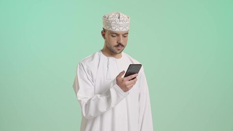 الدردشة مع الأهل والأصدقاء، استخدام جهاز تقني حديث، بورتريه  لشاب عربي خليجي عماني يرتدي الدشداشة و الكمة يمسك بيده الهاتف المحمول، دمج التقنية والتكنولوجيا بالحياة اليومية، تصفح مواقع التواصل الاجتماعي، خلفية خضراء
