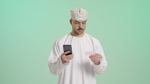 Shopping and payment through mobile programs and applications, modern and advanced technologies, a portrait of a young Arab Gulf Omani man wearing a dishdasha and kumma holding a mobile phone in his hand looking at a credit card with gestures of joy and happiness, green background.