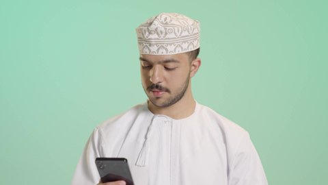 التقنيات الحديثة والمتطورة، الشراء والدفع من خلال برامج وتطبيقات الجوال، بورتريه مقرب لشاب عربي خليجي عماني  يرتدي الدشداشة و الكمة يمسك بيده الهاتف المحمول ينظر الى بطاقة الإئتمان البنكية بإيماءات الفرح و السرور،خلفية خضراء