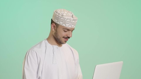 Clicking on the keyboard, the concept of remote business management, using a modern and advanced technical device, a close-up portrait of an Arab Gulf Omani man wearing a dishdasha and kumma using a laptop, integrating technology with work, completing required tasks via the laptop, full-body, green background.