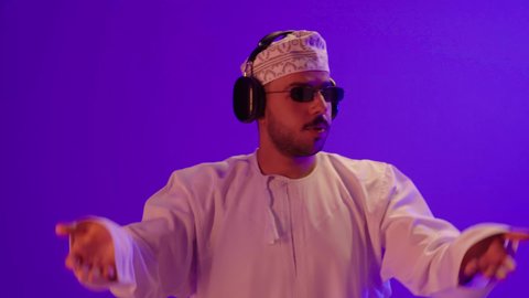 إيماءات وتعابير الاندماج مع الأغنية, مواهب غنائية فنية، بورتريه مقرب لرجل عربي خليجي عماني يرتدي الدشداشه والكمه و النظارة الشسمية يستمع الى الموسيقى من خلال سماعه الرأس، ايماءات تدل على الانصات والإستماع للأغاني بإستخدام الهيدفون، التفاعل مع الانغام الموسيقية، خلفية بنفسجية