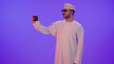 Using the mobile phone to take selfies, making video calls with family and friends, a portrait of a smiling young Arab Gulf Omani man wearing a dishdasha and kumma and sunglasses, holding a mobile phone in his hand and looking at it with gestures of happiness, purple background.