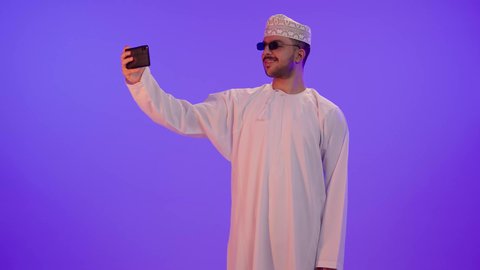 إجراء مكالمات الفيديو مع الأهل والأصدقاء، استخدام الجوال في أخذ لقطات السيلفي، بورتريه لشاب عربي خليجي عماني مبتسم يرتدي الدشداشه والكمة و النظارة الشمسية يمسك الهاتف المحمول بيده وينظر إليه بإيماءات السعادة، خلفية بنفسجية