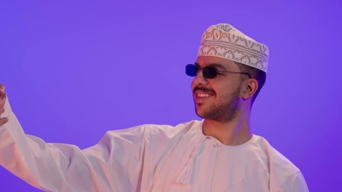 توثيق اللحظات السعيدة, دمج التكنولوجيا والاجهزة الحديثة في الحياة اليومية، بورتريه مقرب لرجل عربي خليجي عماني مبتسم يرتدي الدشداشه والكمة و النظارة الشمسية  يقوم بإلتقاط صور سيلفي تذكارية باستخدام الهاتف المحمول، التواصل مع الاهل والاصدقاء، خلفية بنفسجية