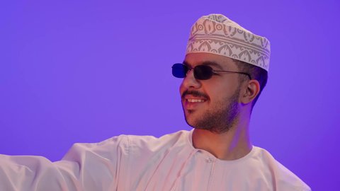 دمج التكنولوجيا والاجهزة الحديثة في الحياة اليومية، بورتريه مقرب لرجل عربي خليجي عماني مبتسم يرتدي الدشداشه والكمة و النظارة الشمسية  يقوم بإلتقاط صور سيلفي تذكارية باستخدام الهاتف المحمول، التواصل مع الاهل والاصدقاء، خلفية بنفسجية
