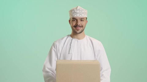 عملية توصيل الطلبات للزبائن، الشحن والتوصيل ذات الجودة العالية، بورتريه لشاب عربي خليجي عماني يرتدي الدشداشة و الكمة يعمل كمندوب مبيعات ويحمل بيده صندوق كرتوني، النظر الى الكاميرا بإيماءات السعادة والسرور، التسوق وطلب المنتجات عبر الإنترنت, خلفية خضراء