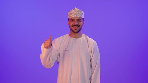 إيماءات الاعجاب والسرور، بورتريه لشاب عربي خليجي عماني مبتسم يرتدي الدشداشة و الكمة يقف و يرفع اصبع الابهام بتعابير الموافقة والرضى والقبول، خلفية بنفسجية