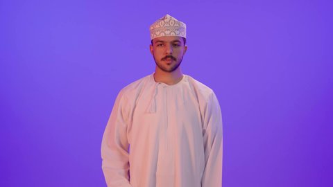 Expressions of agreement, satisfaction, and acceptance, a portrait of a smiling young Arab Gulf Omani man wearing a dishdasha and kumma, standing and raising his thumb up with gestures of admiration and joy, with a purple background.