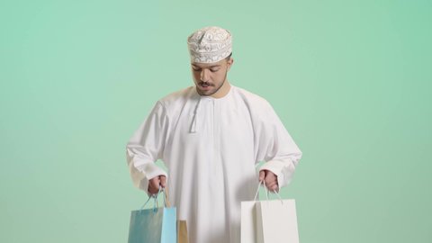 الاستفادة من الحسومات والتصفيات, أكياس الهدايا الورقية التجارية, بورتريه لشاب عربي خليجي عماني يرتدي الدشداشة و الكمة يمسك بيده حقائب المشتريات بايماءات بسعادة, التبضع وأخذ الحاجات الضرورية, متعة التسوق فى المول, خلفية خضراء