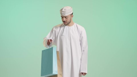 أكياس الهدايا الورقية التجارية، الاستفادة من الحسومات والتصفيات, بورتريه لشاب عربي خليجي عماني يرتدي الدشداشة و الكمة يمسك بيده حقائب المشتريات بايماءات بسعادة, التبضع وأخذ الحاجات الضرورية, متعة التسوق فى المول, خلفية خضراء