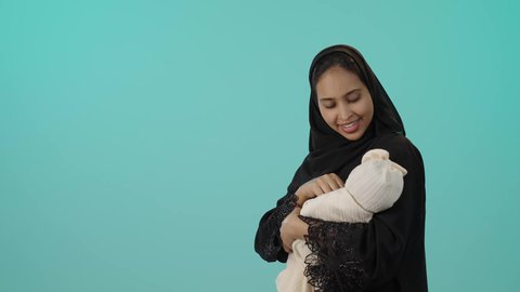 الاستمتاع باللعب مع الأطفال، رعاية الأم لأبنائها والعناية بهم، ايماءات السعادة و السرور، بورتريه لأم عربية خليجية سعودية محجبة تحمل طفلتها الرضيعة بحب وحنان، مفهوم الأمومة والطفولة، خلفية خضراء