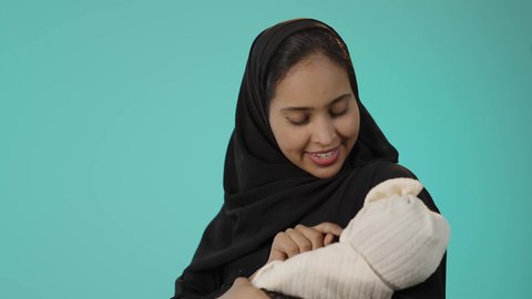 قضاء أوقات ممتعة باللعب مع الأطفال، رعاية الأم لأبنائها والعناية بهم، ايماءات السعادة و السرور، بورتريه مقرب لأم عربية خليجية سعودية محجبة تحمل طفلتها الرضيعة بحب وحنان، مفهوم الأمومة والطفولة، خلفية خضراء