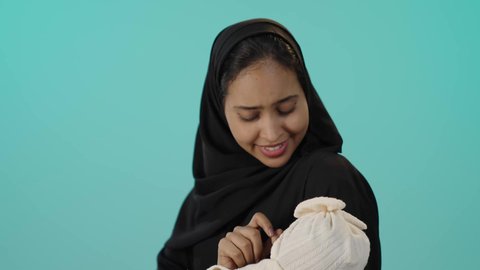 رعاية الأم لأبنائها والعناية بهم، ايماءات السعادة و السرور، بورتريه مقرب لأم عربية خليجية سعودية محجبة تحمل طفلتها الرضيعة بحب وحنان، مفهوم الأمومة والطفولة، قضاء أوقات ممتعة باللعب مع الأطفال، خلفية خضراء