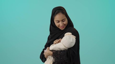 ايماءات السعادة و السرور، رعاية الأم لأبنائها والعناية بهم، بورتريه لأم عربية خليجية سعودية محجبة تحمل طفلتها الرضيعة بحب وحنان، مفهوم الأمومة والطفولة، قضاء أوقات ممتعة باللعب مع الأطفال، خلفية خضراء