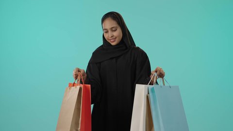 متعة التسوق وانعكاسها على المرأة، الاستمتاع بالتسوق وشراء المستلزمات الجديدة، الترويج للمنتجات والماركات العالمية، بورتريه لشابة عربيه خليجيه سعودية سمراء محجبة ترتدي عباءة تحمل بيدها أكياس تسوق ملونة، النظر للكاميرا بتعابير السعادة، خلفية خضراء