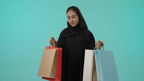 Enjoying shopping and buying new supplies, the positive impact of shopping on women, promoting global products and brands, a portrait of a young Arab Gulf Saudi woman with a dark complexion wearing a hijab and an abaya, holding colorful shopping bags in her hand, looking at the camera with expressions of happiness, green background.