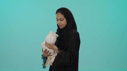 إيماءات الراحة والاسترخاء، الاستمتاع بمشاعر الأمومة، احتضان الطفلة للتهدئة والدخول في النوم العميق، بورتريه لإمرأة عربية خليجية سمراء سعودية ترتدي العباءة تحمل صغيرتها بعطف وعناية، دور الأم الأساسي في رعاية الأبناء، خلفية خضراء