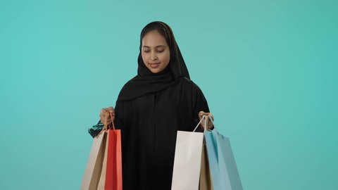 النظر للكاميرا بتعابير السعادة، الترويج للمنتجات والماركات العالمية، الأثر الايجابي للتسوق على المرأة، بورتريه لشابة عربيه خليجيه سعودية سمراء محجبة ترتدي عباءة تحمل بيدها أكياس تسوق ملونة، الاستمتاع بالتسوق وشراء المستلزمات الجديدة، خلفية خضراء