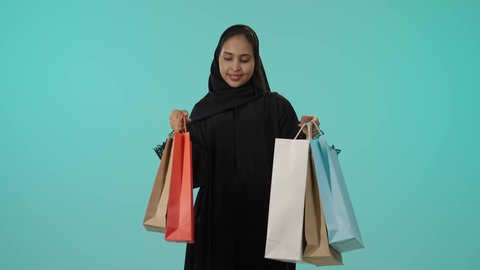 الترويج للمنتجات والماركات العالمية، الأثر الايجابي للتسوق على المرأة، بورتريه لشابة عربيه خليجيه سعودية سمراء محجبة ترتدي عباءة تحمل بيدها أكياس تسوق ملونة، النظر للكاميرا بتعابير السعادة، الاستمتاع بالتسوق وشراء المستلزمات الجديدة، خلفية خضراء