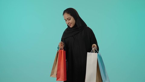 الأثر الايجابي للتسوق على المرأة، الترويج للمنتجات والماركات العالمية،  بورتريه لشابة عربيه خليجيه سعودية سمراء محجبة ترتدي عباءة تحمل بيدها أكياس تسوق ملونة، النظر للكاميرا بتعابير السعادة، الاستمتاع بالتسوق وشراء المستلزمات الجديدة، خلفية خضراء