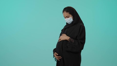 الوقاية من فايروس كورونا، الحفاظ على صحتها و صحة طفلها، الاهتمام بصحة الجنين، مفهوم الحمل والانجاب، بورترية لامراة عربية خليجية سعودية ترتدي العباءة السوداء و الكمامة تضع يدها على بطنها تنتظر قدوم مولودها، النظر إلى الكاميرا بإيماءات الحماس، خلفية خضراء