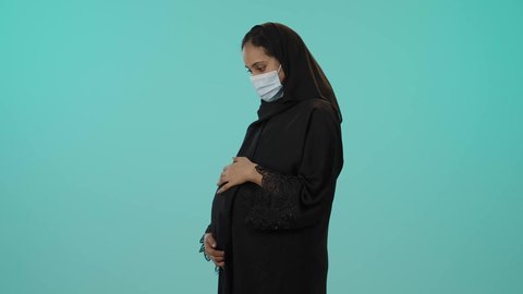 مفهوم الحمل والانجاب، الحفاظ على صحتها و صحة طفلها، الاهتمام بصحة الجنين، بورترية لامراة عربية خليجية سعودية ترتدي العباءة السوداء و الكمامة تضع يدها على بطنها تنتظر قدوم مولودها، النظر إلى الكاميرا بإيماءات الحماس، الوقاية من فايروس كورونا، خلفية خضراء