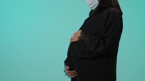 الاهتمام بصحة الجنين، الحفاظ على صحتها و صحة طفلها، مفهوم الحمل والانجاب، بورترية مقرب لامراة عربية خليجية سعودية ترتدي العباءة السوداء و الكمامة تضع يدها على بطنها تنتظر قدوم مولودها، النظر إلى الكاميرا بإيماءات الحماس، الوقاية من فايروس كورونا، خلفية خضراء