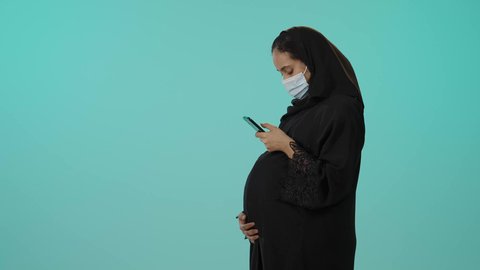 تصفح تطبيقات الهاتف المحمول، الوقاية من فايروس كورونا، الحفاظ على صحتها و صحة طفلها، الاهتمام بصحة الجنين، مفهوم الحمل والانجاب، بورترية لامراة عربية خليجية سعودية ترتدي العباءة السوداء و الكمامة تمسك الجوال وتضع يدها على بطنها تنتظر قدوم مولودها، خلفية خضراء