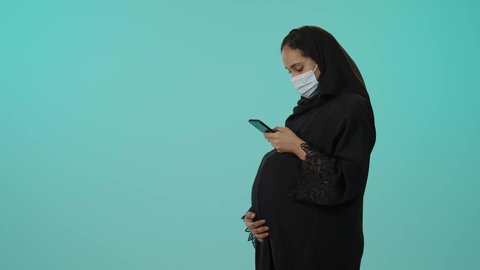 متابعة اخبار وسائل التواصل الاجتماعي باستخدام الجوال، الوقاية من فايروس كورونا، الحفاظ على صحتها و صحة طفلها، الاهتمام بصحة الجنين، مفهوم الحمل والانجاب، بورترية لامراة عربية خليجية سعودية ترتدي العباءة السوداء و الكمامة تمسك الهاتف المحمول وتضع يدها على بطنها تنتظر قدوم مولودها، خلفية خضراء