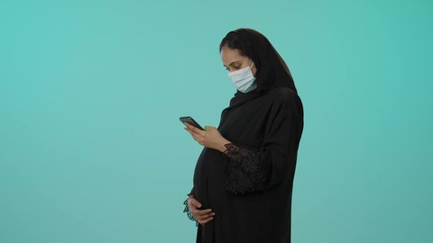 الدردشة والتحدث مع الاصدقاء عبر الانترنت، الوقاية من فايروس كورونا، الحفاظ على صحتها و صحة طفلها، الاهتمام بصحة الجنين، مفهوم الحمل والانجاب، بورترية لامراة عربية خليجية سعودية ترتدي العباءة السوداء و الكمامة تمسك الهاتف المحمول وتضع يدها على بطنها تنتظر قدوم مولودها، خلفية خضراء