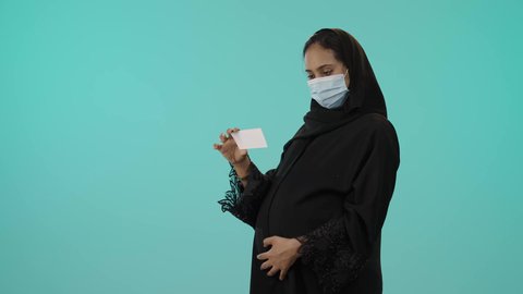 إظهار نتيجة السونار في ورقة، مفهوم الحمل والأمومة، تصوير لامراة عربيه خليجيه سعودية محجبه ترتدي العباءة السوداء و الكمامة تقوم بعرض صورة أشعة لجنين، الوقاية من فايروس كورونا ، خلفية خضراء
