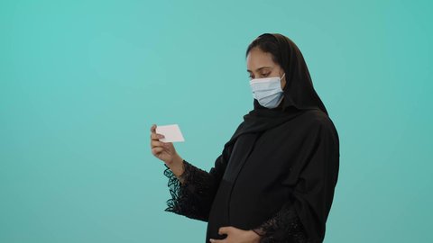 صورة أشعة بالموجات فوق الصوتية، مفهوم الحمل والأمومة، تصوير لامراة عربيه خليجيه سعودية محجبه ترتدي العباءة السوداء و الكمامة تقوم بعرض صورة أشعة لجنين، الوقاية من فايروس كورونا ، خلفية خضراء