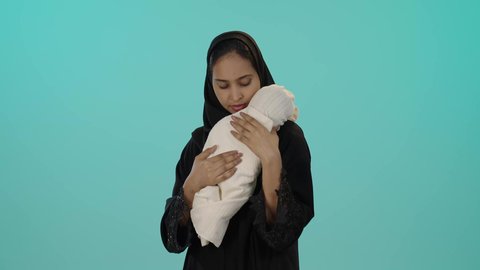 الأسرة السعودية وتحمل المسؤولية, حنان الأمهات و رعاية الأبناء، بورتريه لأم عربية خليجية سعودية محجبة تحمل رضيعتها بحب وحنان و تقوم بتقبيلها، إيماءات السعادة والأمان، مفهوم الأمومة والطفولة، قضاء أوقات ممتعة بالعناية بالأطفال، خلفية خضراء