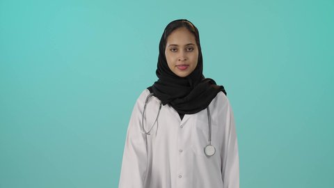 Quality and excellence mark, the concept of admiration and satisfaction, medical care for patients, a portrait of a dark-skinned Saudi Gulf Arab female doctor wearing a white medical coat, pointing her thumb up in gestures of approval, with a green background.
