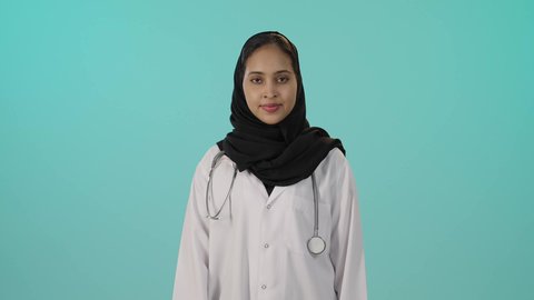 مفهوم الإعجاب والرضا، علامة الجودة و الامتياز, الرعاية الطبية للمرضى، بورتريه لطبيبة عربية خليجية سعودية سمراء ترتدي المعطف الطبي الأبيض تشير بإصبع الابهام الى الاعلى بإيماءات الموافقة، خلفية خضراء