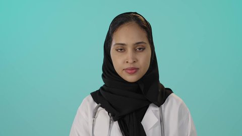 الوقوف باستقامة و اعتدال, حركات الرفض و الامتناع, بورتريه مقرب لطبيبة عربية خليجية سعودية سمراء محجبه ترتدي المعطف الطبي و السماعات تنظر إلى الكاميرا بتعابير الجدية و الحزم,إيماءات طلب التوقف و عدم الاقتراب, خلفية خضراء