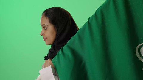 ذكرى العيد الوطني السعودي 23 سبتمبر، تأسيس الدولة السعودية الأولى 22 فبراير، ذكرى يوم بدينا 1727 م, بورتريه من الجانب لشابة عربية خليجية سعودية سمراء تحمل علم المملكه على ظهرها  بتعابير الفخر والاعتزاز، خلفية خضراء